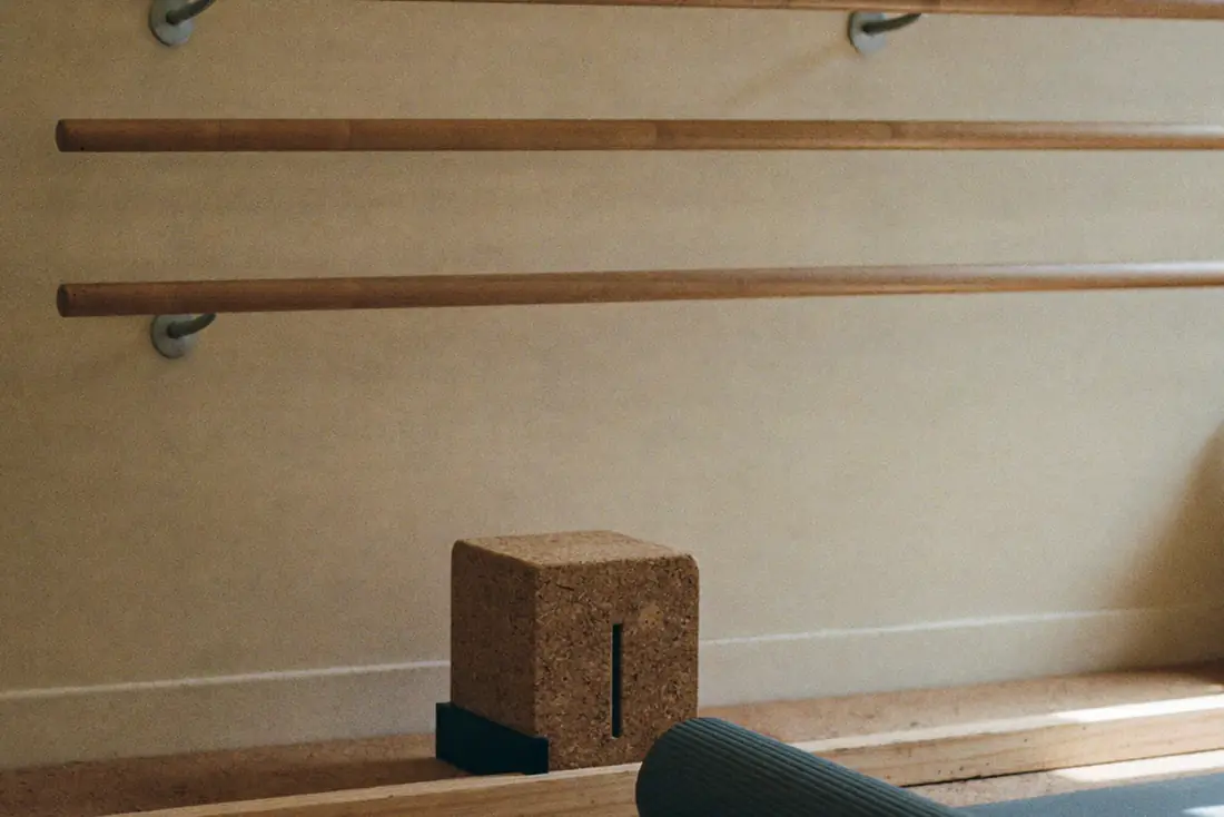 A wall-mounted ash wood handrail and a low wooden balance beam on a cork floor, with a rolled slate blue mat and soft window light across the wood