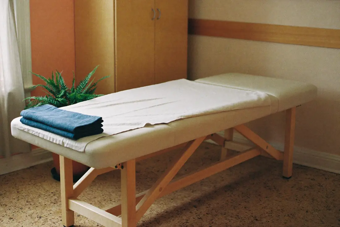 An empty treatment room in morning light: a padded table with a linen cover and a folded slate blue towel, a pale wood cabinet, a sheer curtain and a fern