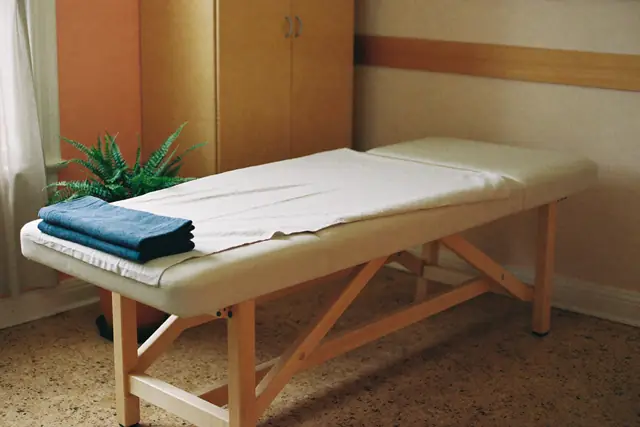 An empty treatment room in morning light: a padded table with a linen cover and a folded slate blue towel, a pale wood cabinet, a sheer curtain and a fern